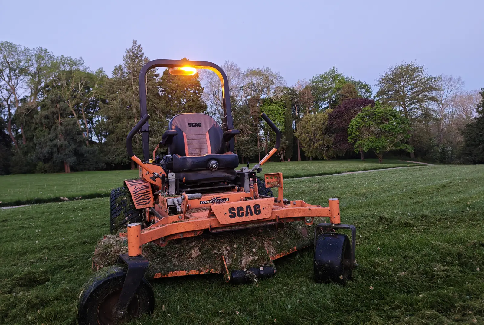 Orange SCAG ride-on mower with its work light on, parked on a freshly mown park lawn at dusk
