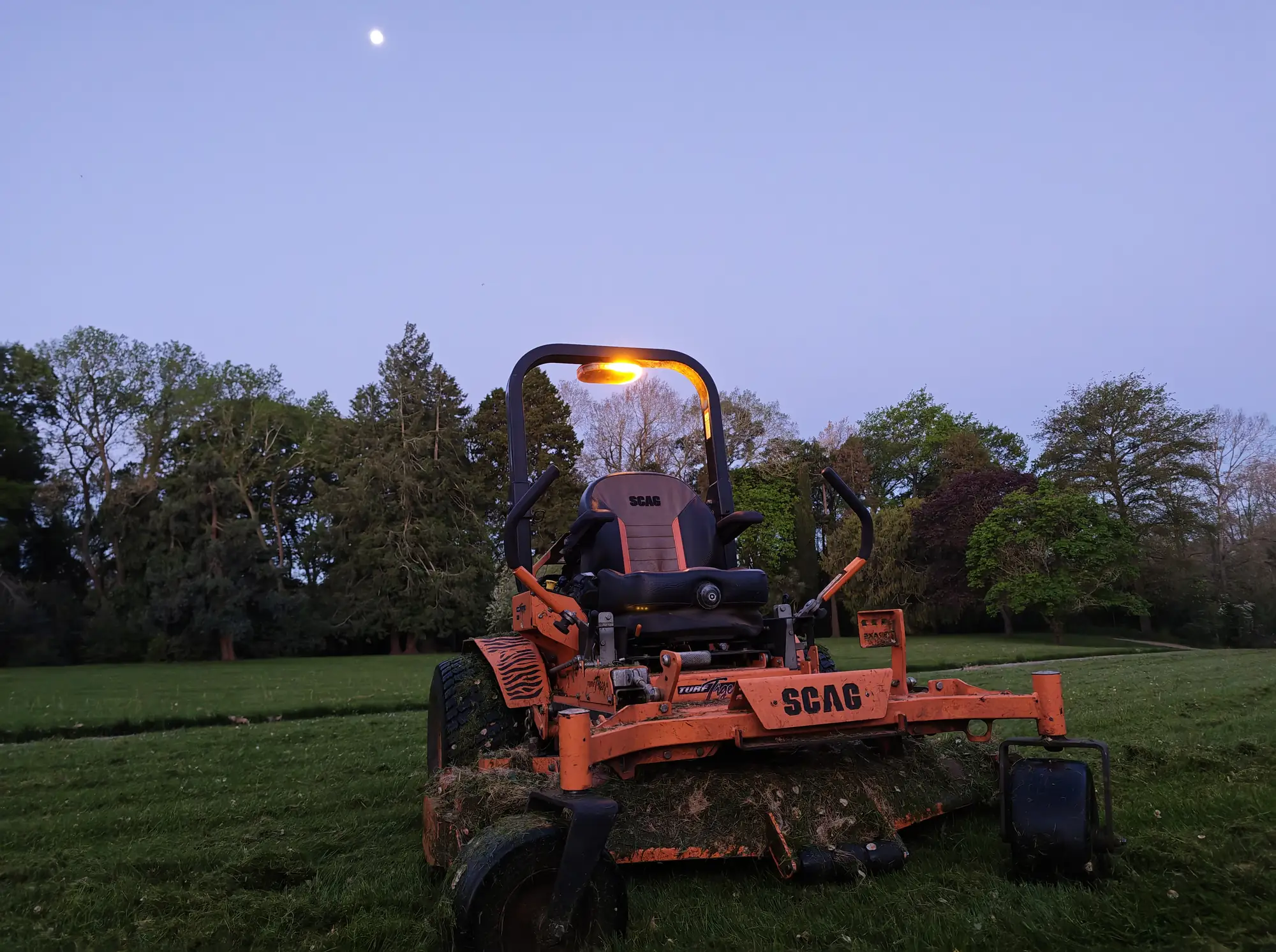 Orange SCAG ride-on mower on a freshly mown park lawn at dusk, with the moon above the trees